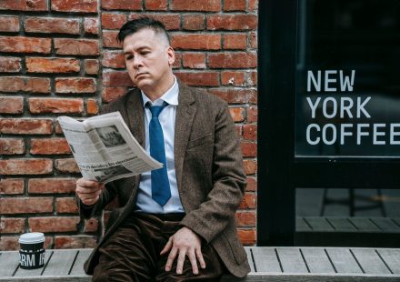 A professional man in formal wear reads a newspaper beside a coffee shop window. — test123123